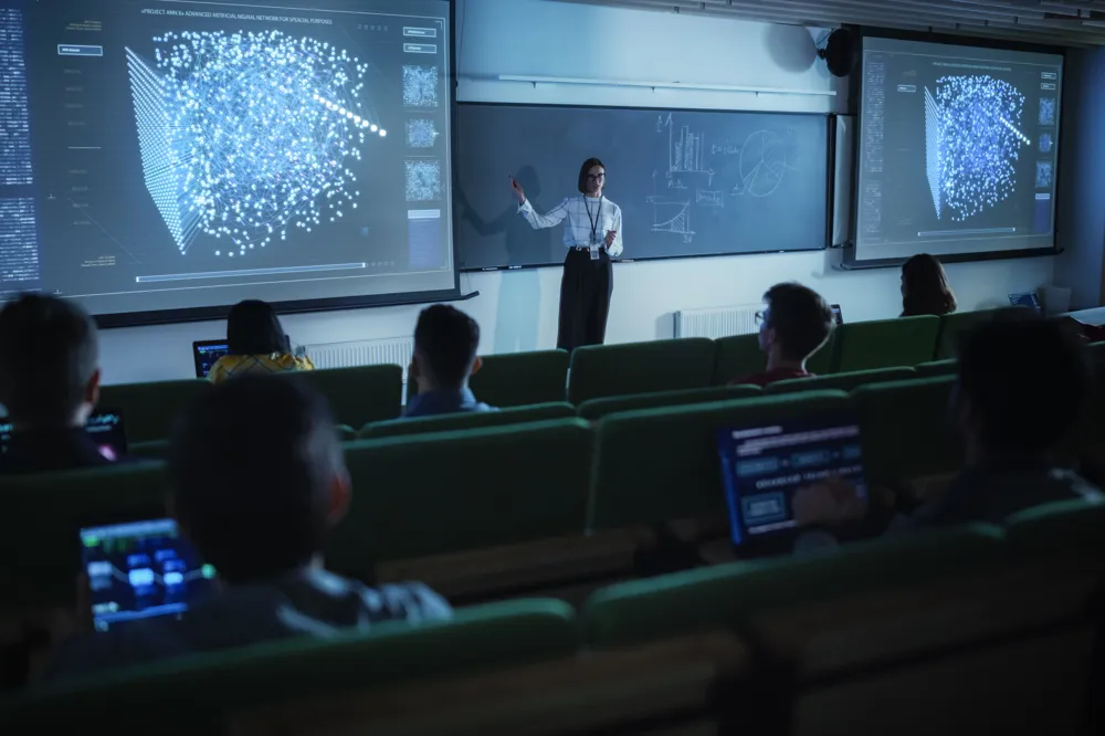 ALT text: A female presenter in a tech-focused classroom explains a 3D quantum displayed on two large screens, addressing students seated with laptops.